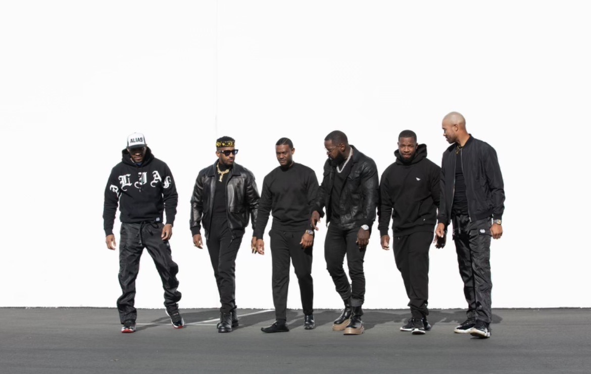 A group of six men in dark clothing standing together against a white backdrop