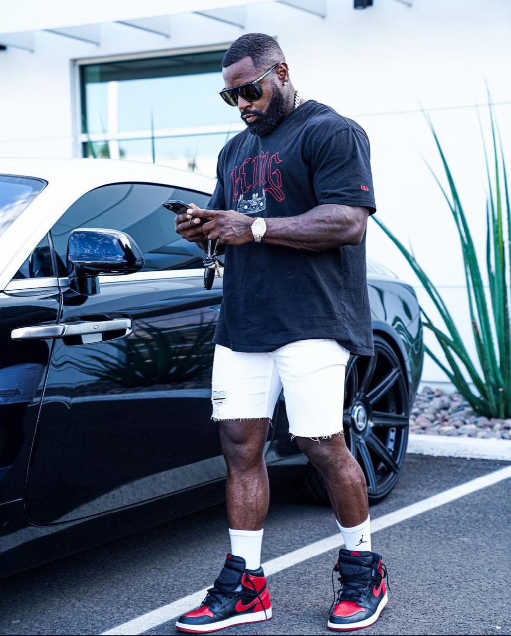 Mike Rashid checking his phone beside a dark car, wearing a King shirt and red sneakers