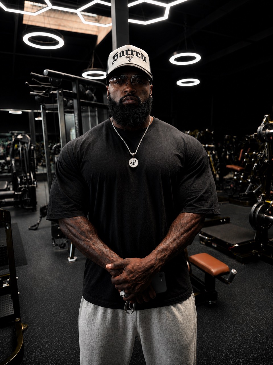 Mike Rashid King in a black shirt and Sacred cap, standing in a gym beneath ring lights